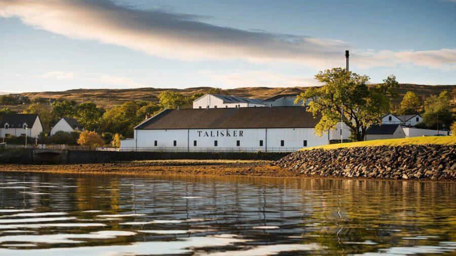 Talisker Distillery on Isle of Skye, Scotland, by tranquil loch and lush green hills.
