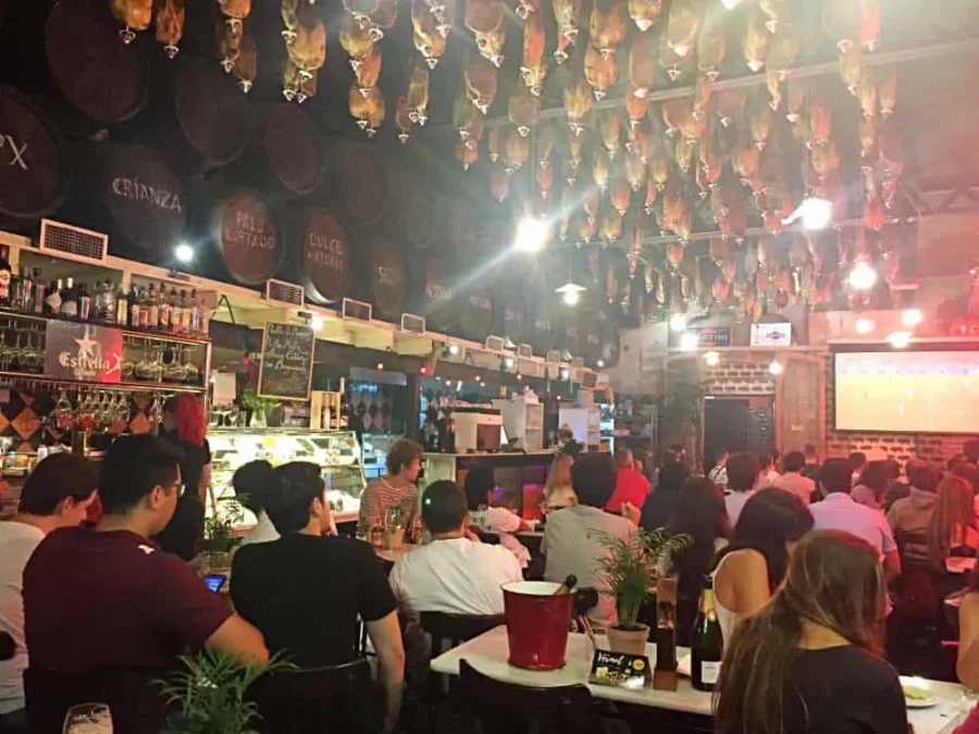 Crowded tapas bar with hanging cured hams, wine barrels, warm lighting, and patrons watching sports.