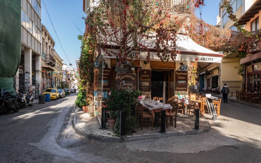 Psyrri Athens Greek taverna on a sunny corner street with outdoor seating and vine-covered awning.