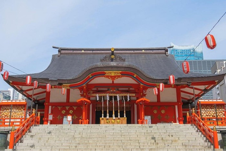 Hanazono Shinto shrine with stone steps, vermilion facade, lanterns, and urban skyline.