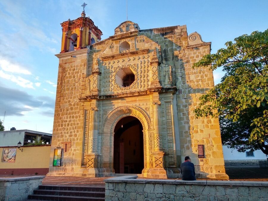 Templo de San Matías Jalatlaco features a prominent tower, with a man resting on the steps in front of the church.
