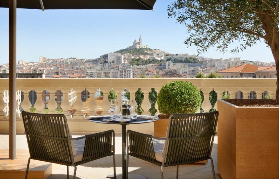 Outdoor terrace with cityscape view, modern chairs, table setting, plants, and iconic structure.