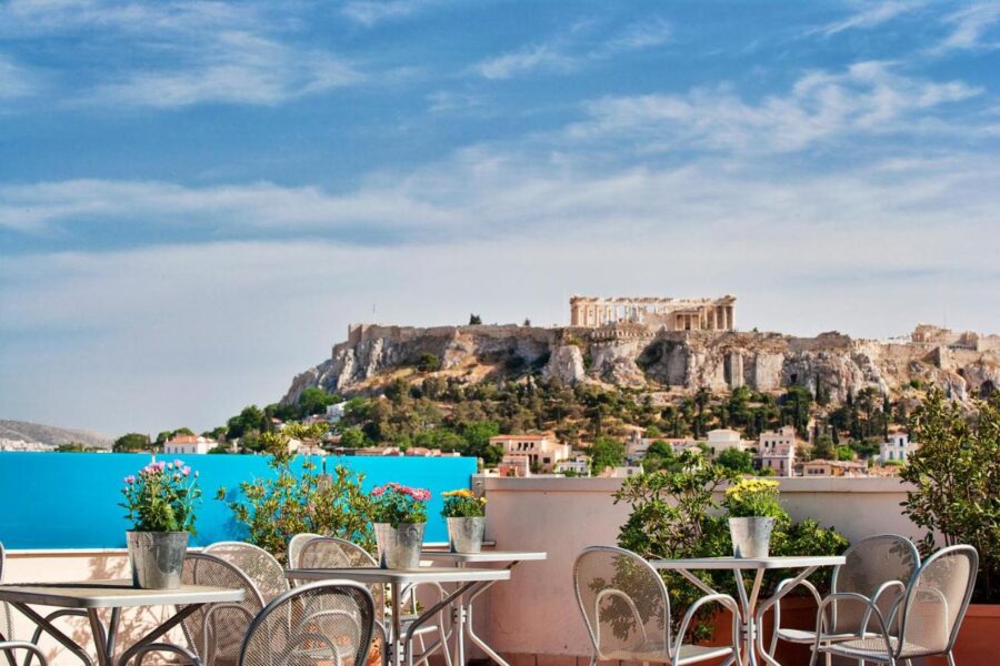 Rooftop terrace in Athens with tables, plants, and Acropolis view under blue sky.