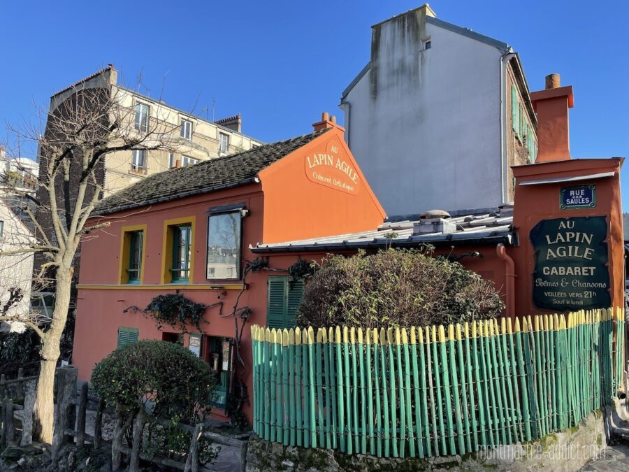 Terracotta cabaret Au Lapin Agile on Rue Saules with green shutters and turquoise picket fence.