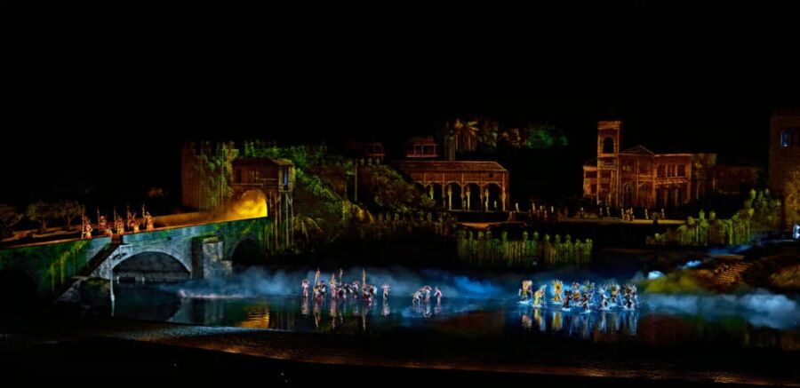 El Sueno de Toledo Show Dancers in vibrant costumes perform on water amidst illuminated, ancient-style buildings, exuding a mystical and otherworldly atmosphere.
