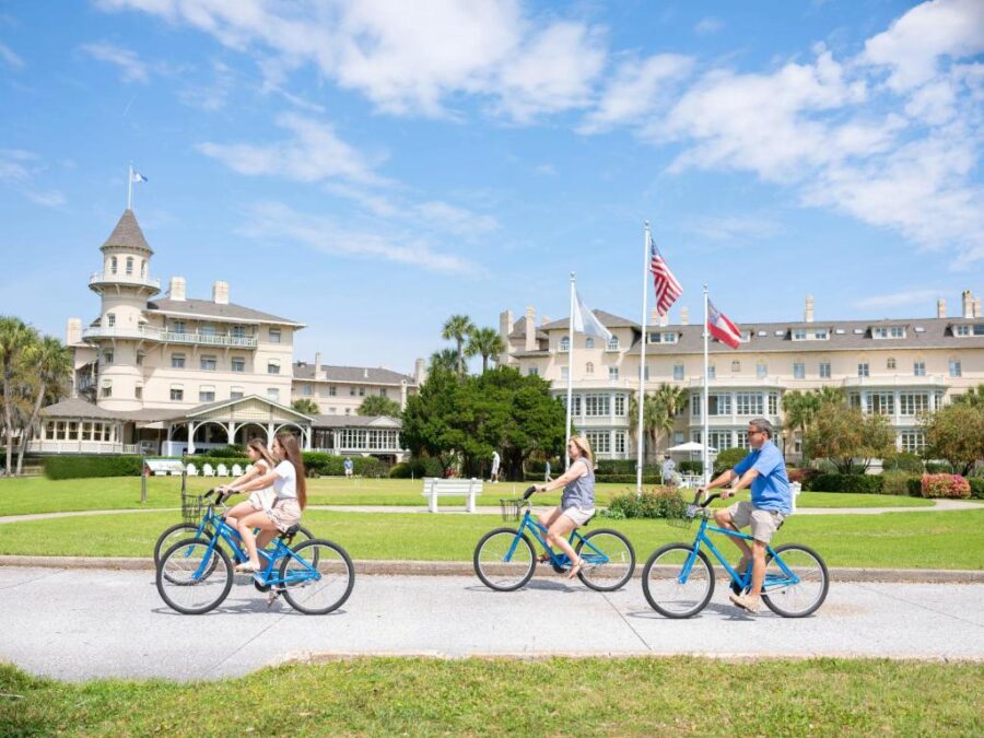 Three people bike past the grand white Jekyll Island Club Resort in Georgia, showcasing the scenic surroundings.
