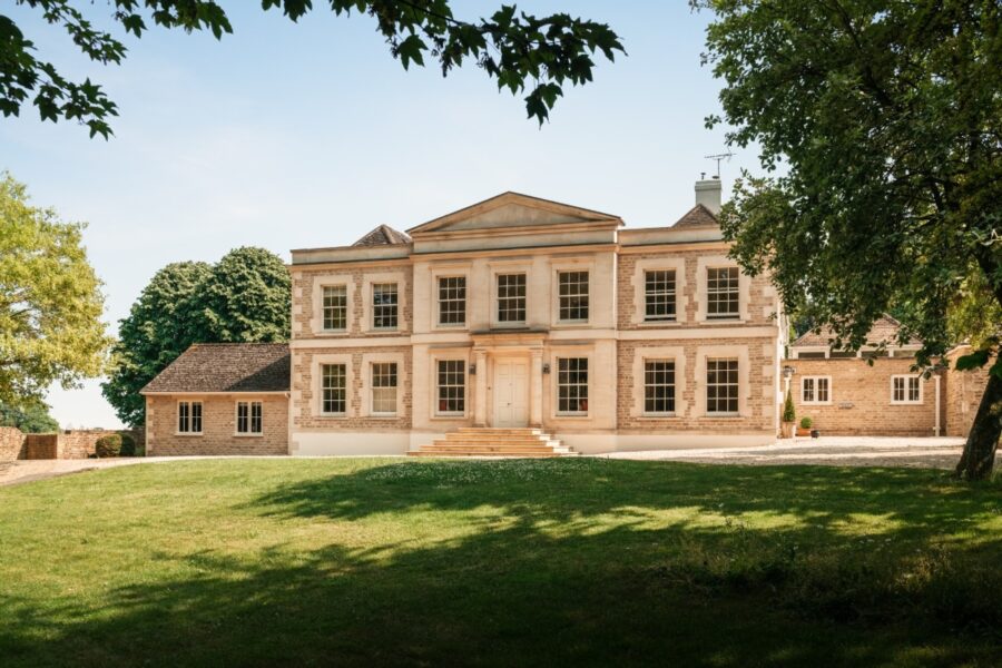 The Palladian house in the Cotswold, UK, is enveloped by trees and a grassy landscape.