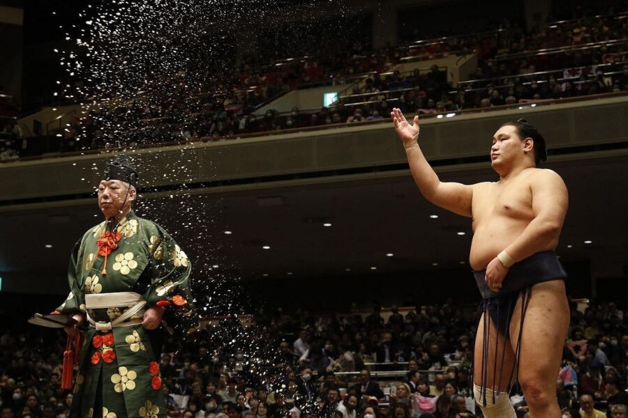 Sumo wrestler performs a ceremonial salt toss in a crowded arena. Beside him, a referee stands in traditional attire. The scene is tense and traditional.