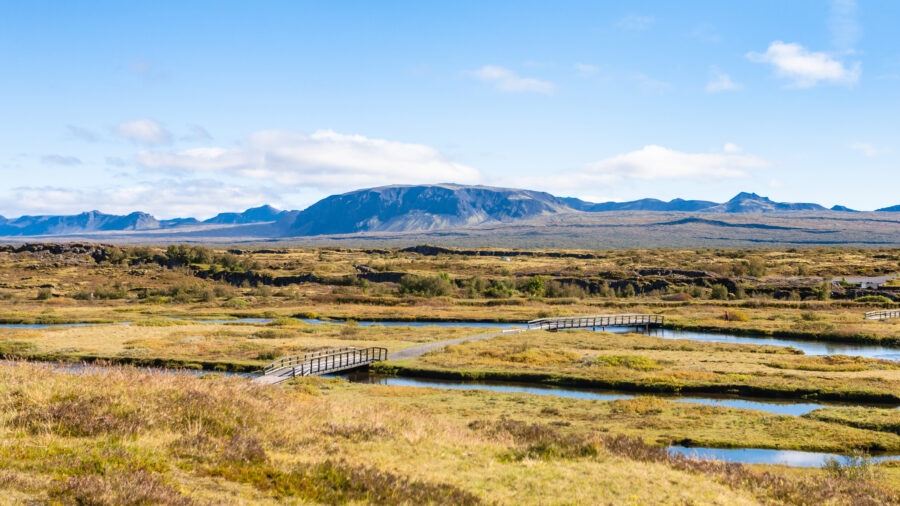 Expansive landscape of Silfra Fissure Thingvellir Park with green fields and winding streams in the foreground. A wooden footbridge spans the water. Mountains under a bright blue sky.