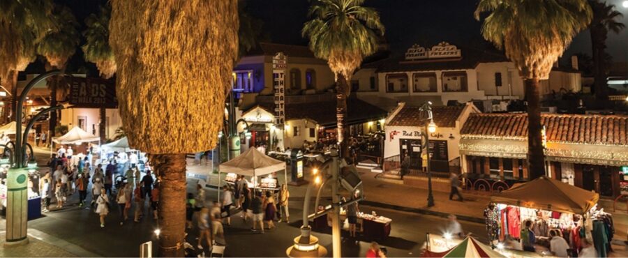 Night view of a lively street at Thursday VillageFest, featuring palm trees and people enjoying the festive atmosphere.
