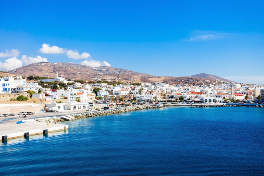 Tinos Island coastal town with white buildings and colorful roofs beside a clear blue sea. Hills in the background under a bright sky. Serene and picturesque.