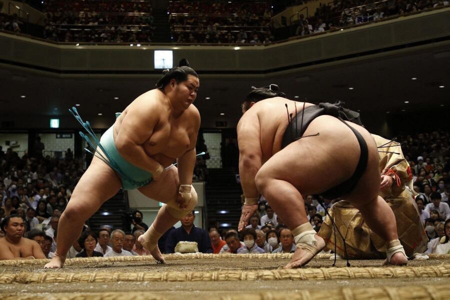 Two sumo wrestlers crouch in a ring, poised to engage, in front of an audience in a large arena. The crowd watches intently, creating a tense atmosphere.