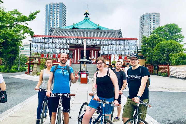 Group of five people with bicycles smile in front of a traditional Japanese temple in Tokyo. Skyscrapers and lush greenery are visible in the background.