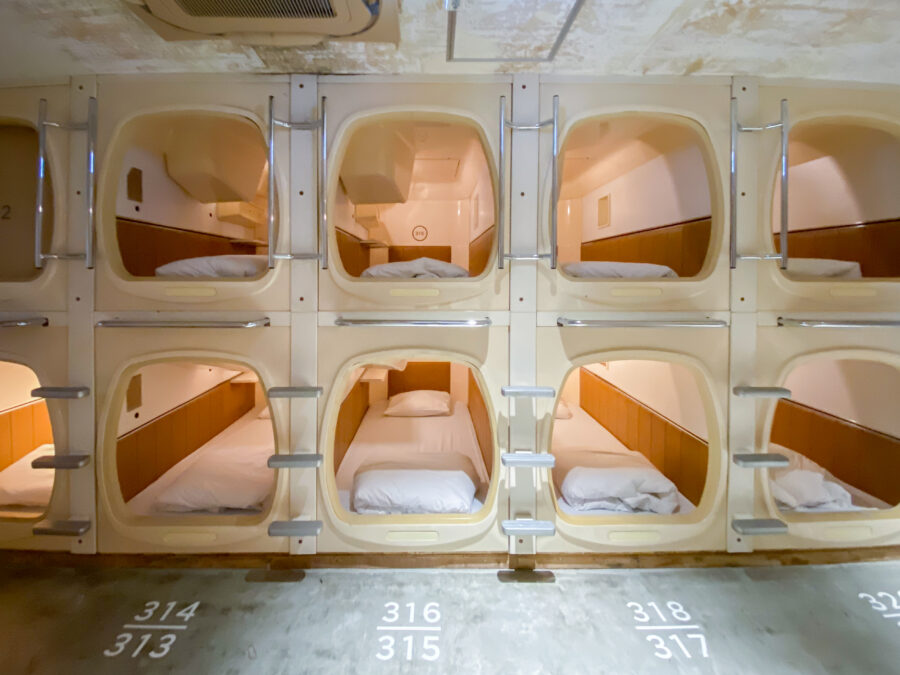 Row of cozy, stacked capsule hotel pods, each with a neatly made bed, pillow, and soft lighting, conveying a futuristic and compact vibe.