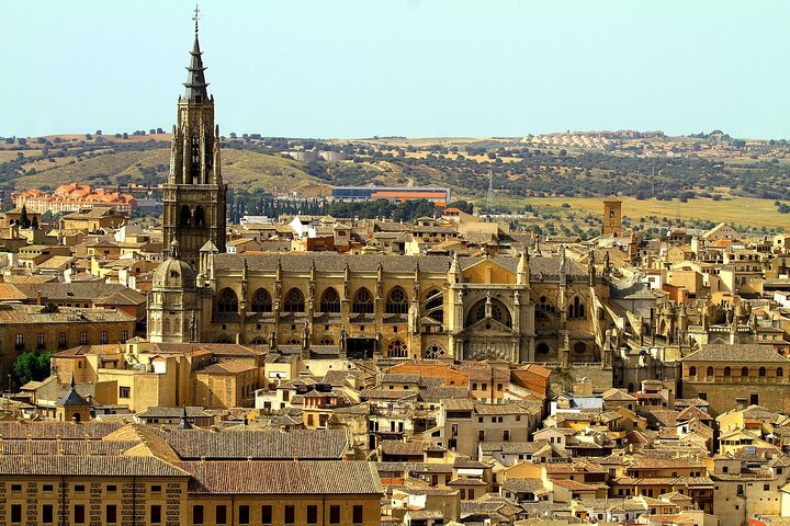 Panoramic view of Toledo, Spain, featuring the Gothic Toledo Cathedral with its tall spire. Historic buildings and rolling hills frame the scene.