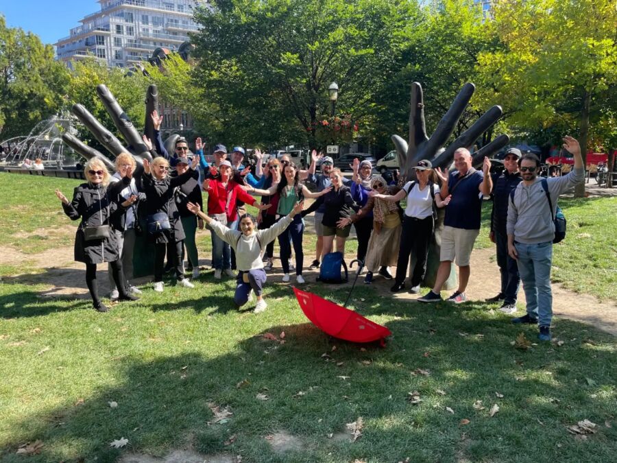 Group of people joyfully pose with raised arms in front of large hand sculptures in a park. A red umbrella lies on the grass, and trees surround the scene.