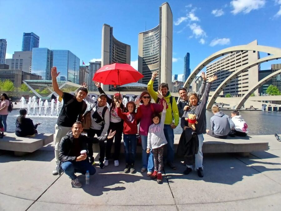 Joyful group of people stands in front of a fountain and modern buildings, waving and smiling on a sunny day. One holds a red umbrella.