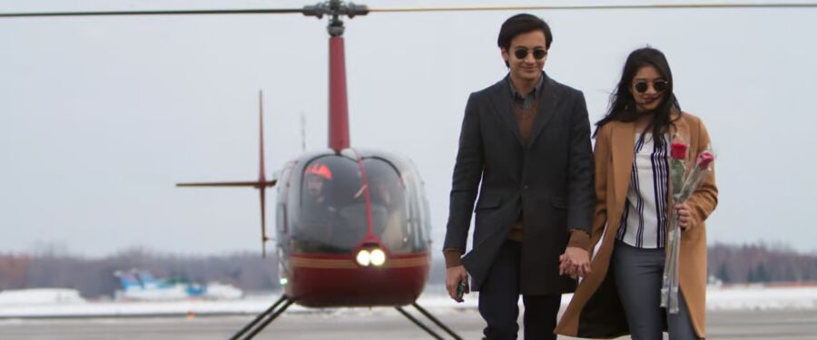 Couple walks hand-in-hand away from a parked helicopter on a runway. The woman holds roses, and both wear sunglasses, exuding sophistication and joy.