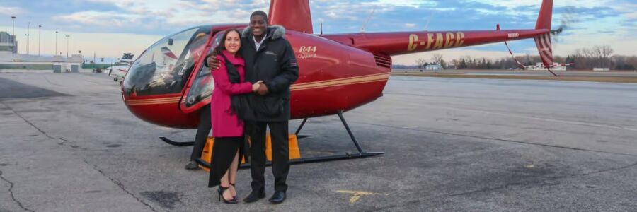 Smiling couple stands in front of a red helicopter on a tarmac. The sky is cloudy, adding depth to the scene. They appear joyful and excited.