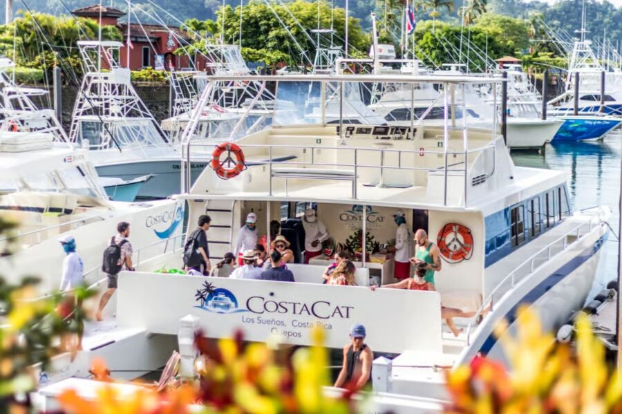 Large catamaran labeled "CostaCat" is docked, with people on board, some wearing hats and backpacks for a Tortuga Beach Day Trip.