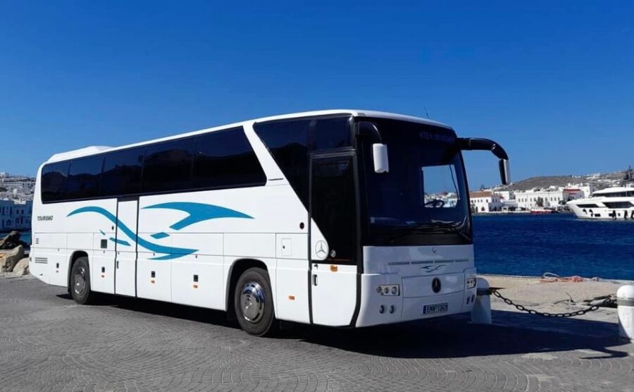 Modern charter bus by scenic coastal waterfront with boats and traditional architecture.
