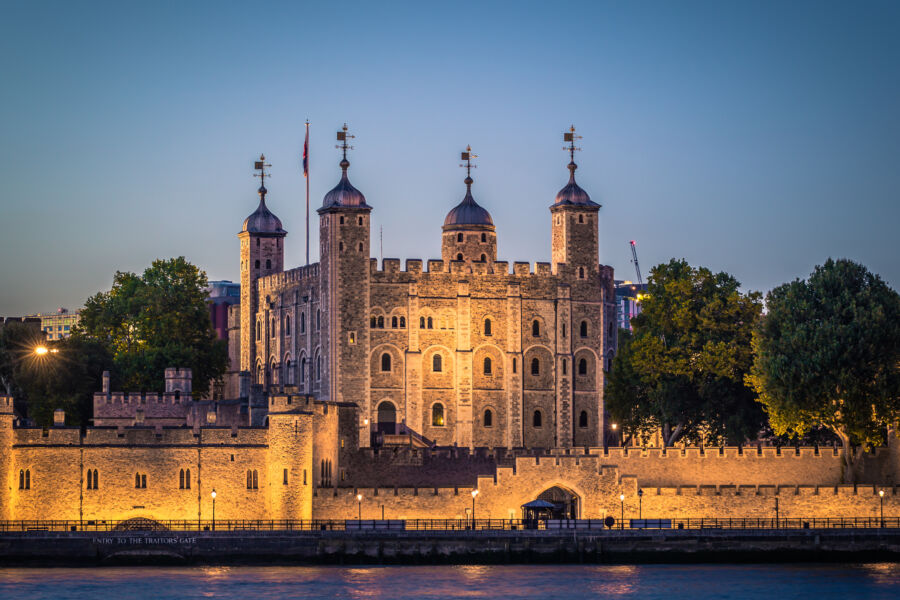 Night view of the Tower of London beside the River Thames, showcasing its historic architecture against a dark sky