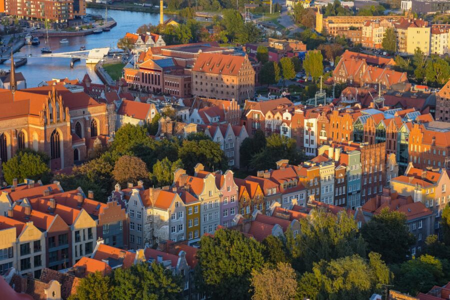 Gdansk urban landscape with Gothic church, rooftops, river, and lush trees in warm light.