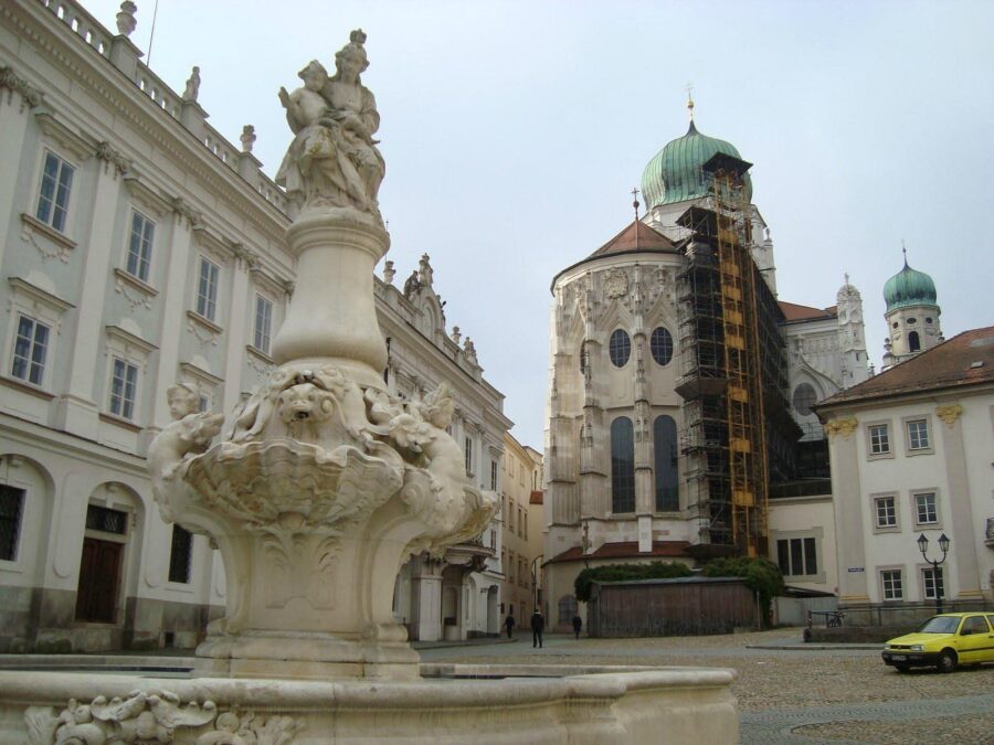 Historic town square with ornate stone fountain and church with green domes on cobblestone plaza.