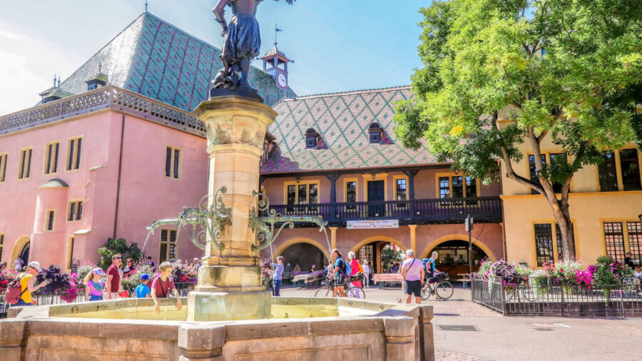 Historic town square with fountain, patterned tiled-roof building, clock tower, and flower beds.