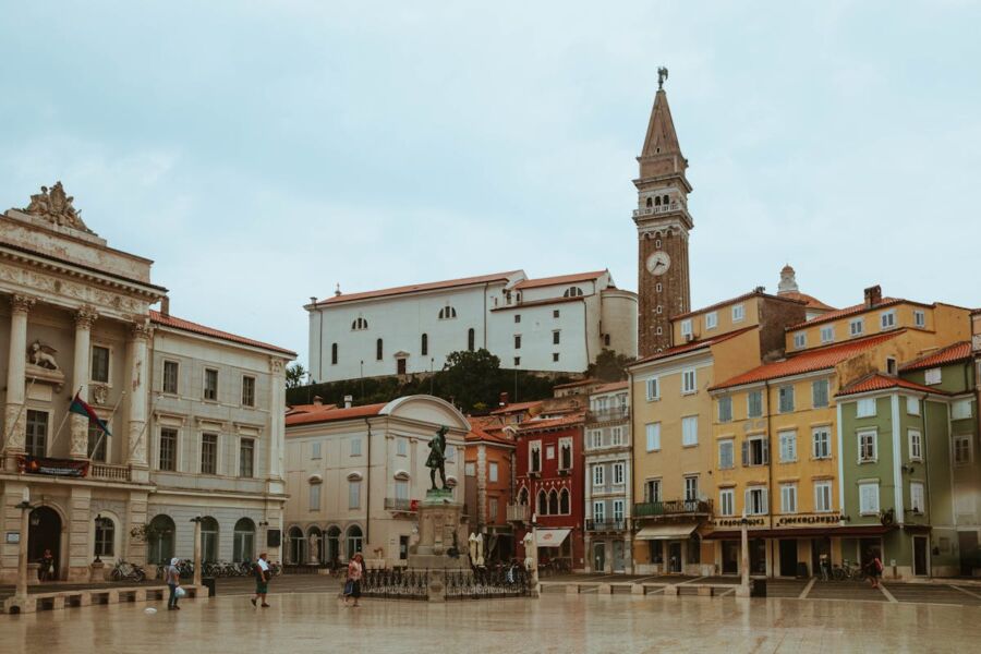 Charming coastal town square with colorful buildings, historic architecture, clock tower, and central statue.