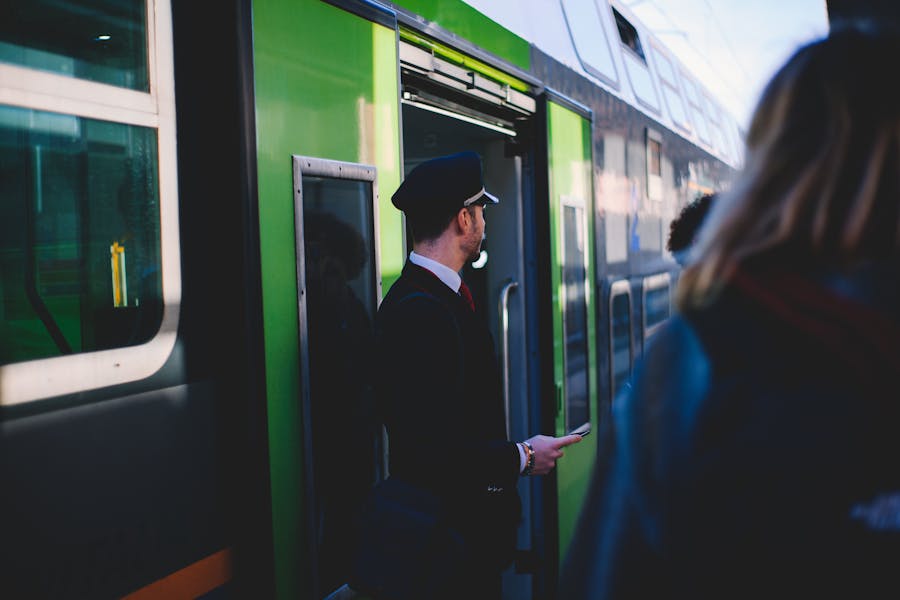 Train conductor in uniform at station with vibrant green train in sunlight.