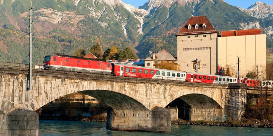 Train crossing stone arch bridge in lush mountain landscape with river and quaint buildings.