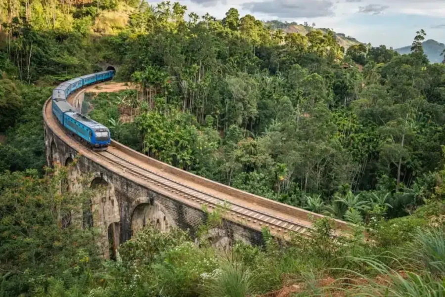 Blue train on stone viaduct amidst lush greenery and rolling hills in a tropical landscape.