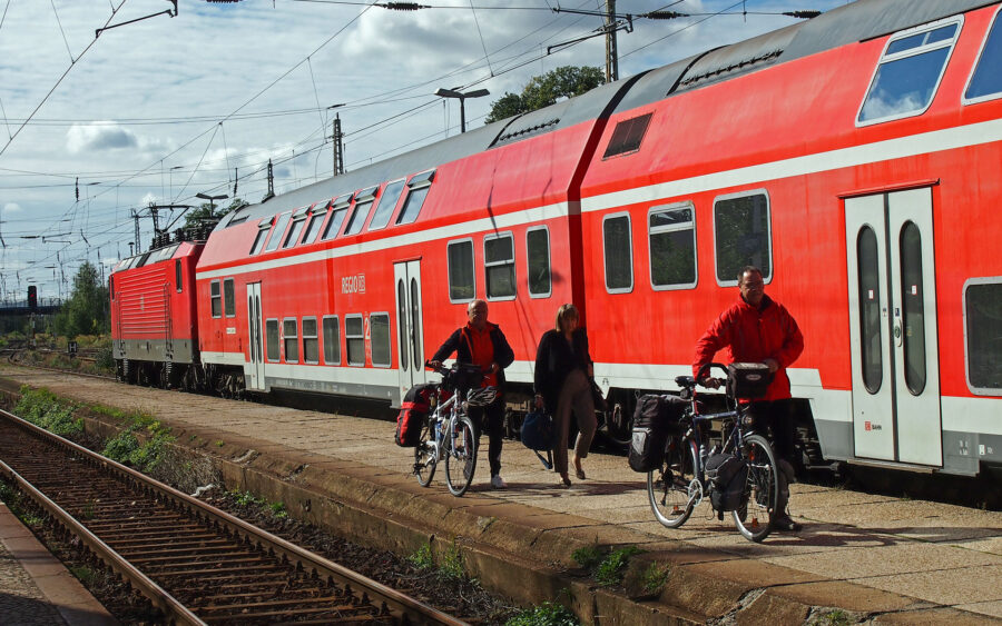 Red double-decker train at station with cyclists and commuters amid urban and natural scenery.