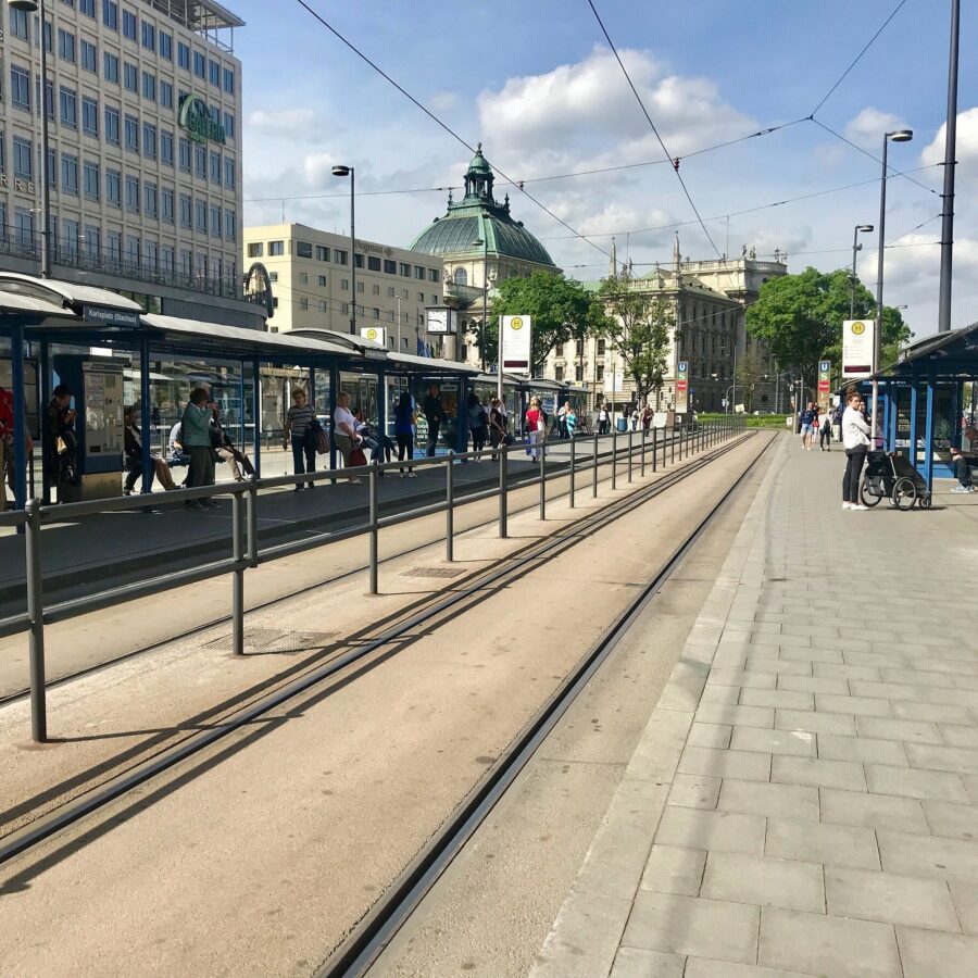 Munich tram station platforms with parallel tracks, commuters, shelters, and historic domed building.