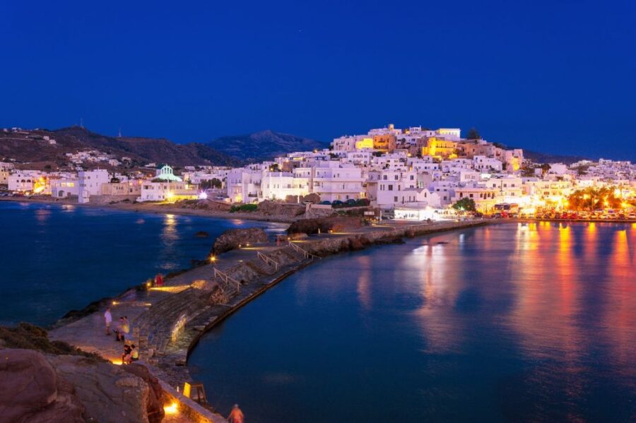 Twilight view of Greek coastal town with promenade, whitewashed buildings, and mountains.