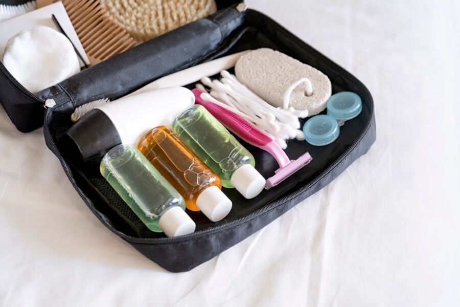 Open black toiletry bag on a white surface containing travel-sized bottles, razor, cotton pads, cotton swabs, pumice stone, and contact lens cases