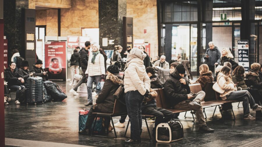 Travelers in a busy train station with winter attire and various luggage.