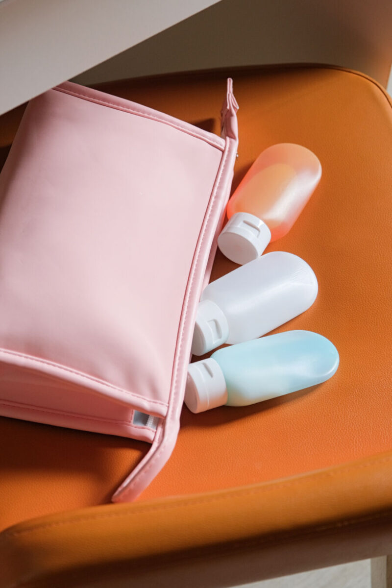 Travel-sized plastic toiletry containers and a pink cosmetic bag on a chair in a hotel room.

