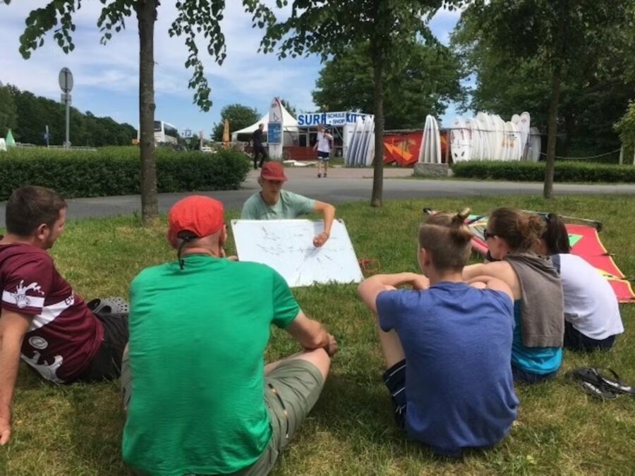 Participants seated on grass in front of a large whiteboard during the Trial Windsurf Course in Zingst.

