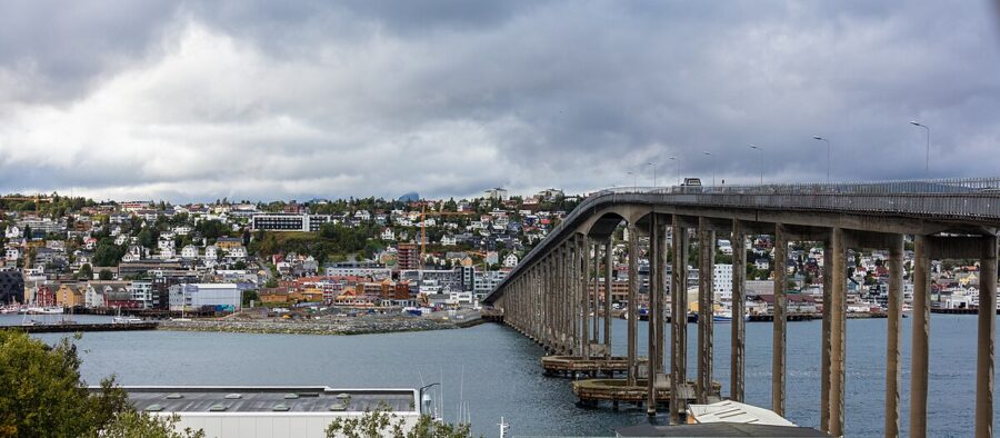 Tromsø Bridge spans over water with the city of Tromsø, Norway, visible in the background under a clear blue sky.