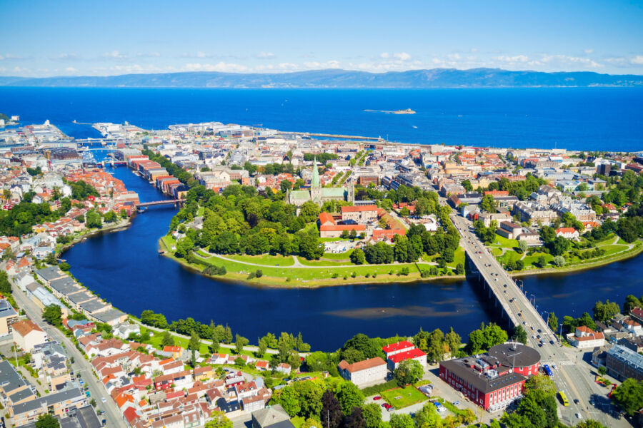 Aerial panoramic view of Trondheim, showcasing the cityscape, river, and surrounding hills under a clear blue sky.
