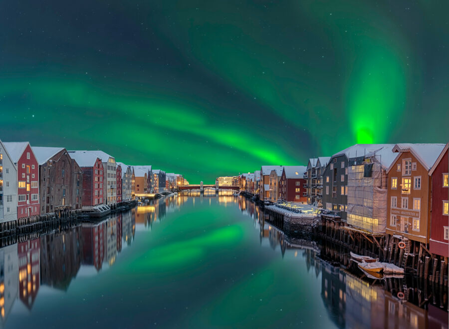 Winter scene in Trondheim with colorful northern lights over the river and traditional wooden houses along the banks.