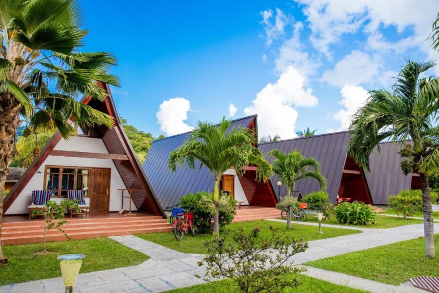 Tropical A-frame bungalows resort with lush palm gardens, wooden decks, and bicycles under blue sky.