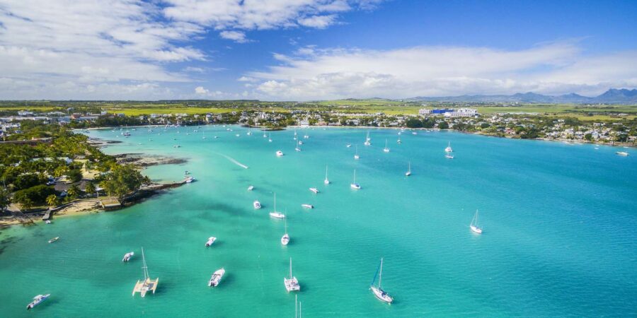 Aerial view of tropical bay with turquoise water, sailboats, palm trees, beach houses and mountains.