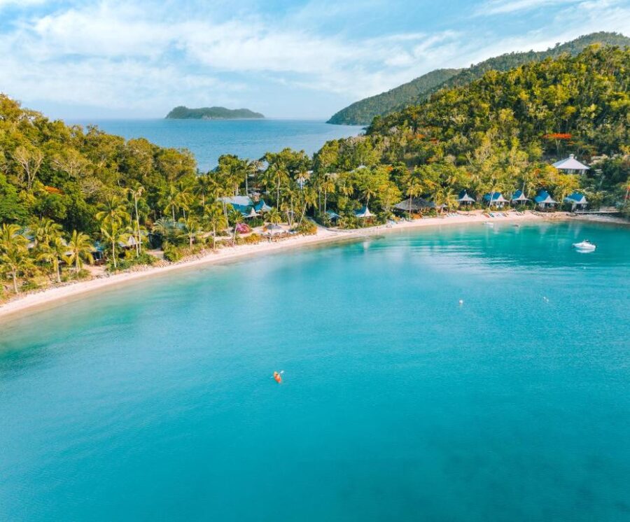 Aerial view of tropical bay resort with turquoise water, sandy beach, palm trees, and kayaker.