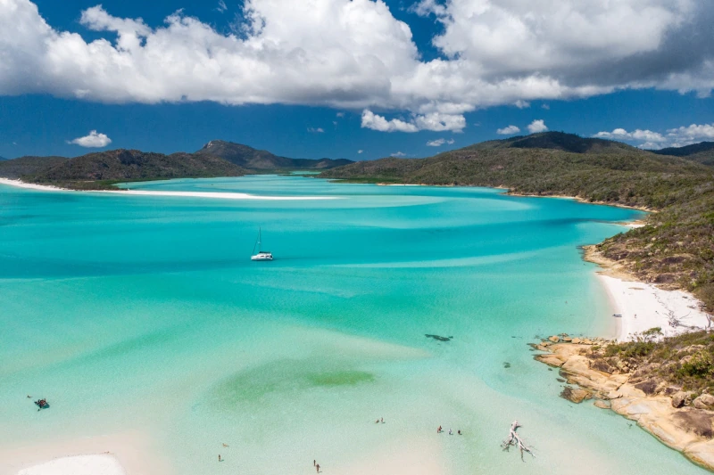 Aerial view of tropical Whitsunday bay with turquoise water, white sand beach, sailboat, and hills.