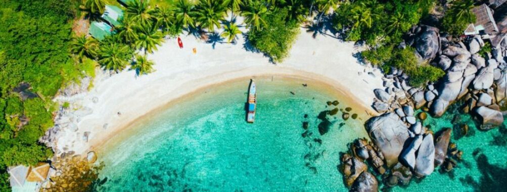 Aerial view of a tropical beach with palm trees, turquoise waters, and a small anchored boat.