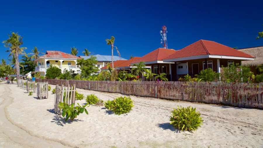 Sunlit tropical beach with sand, palm trees, and beachfront bungalows under a clear blue sky.