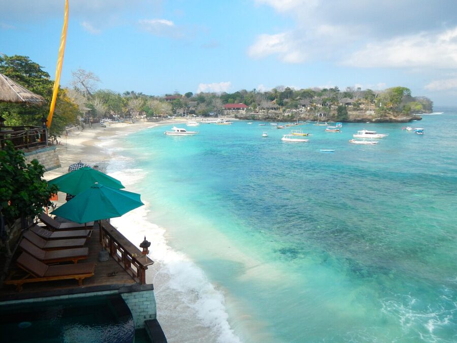 Tropical beach resort terrace overlooking turquoise bay with boats, white sand, and coastal village.